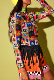 A model wearing a vibrant, multicolored patchwork long-sleeve top with cartoon and animal prints, paired with a black skirt featuring orange flames and pink lace trim, set against a solid yellow-green background.
