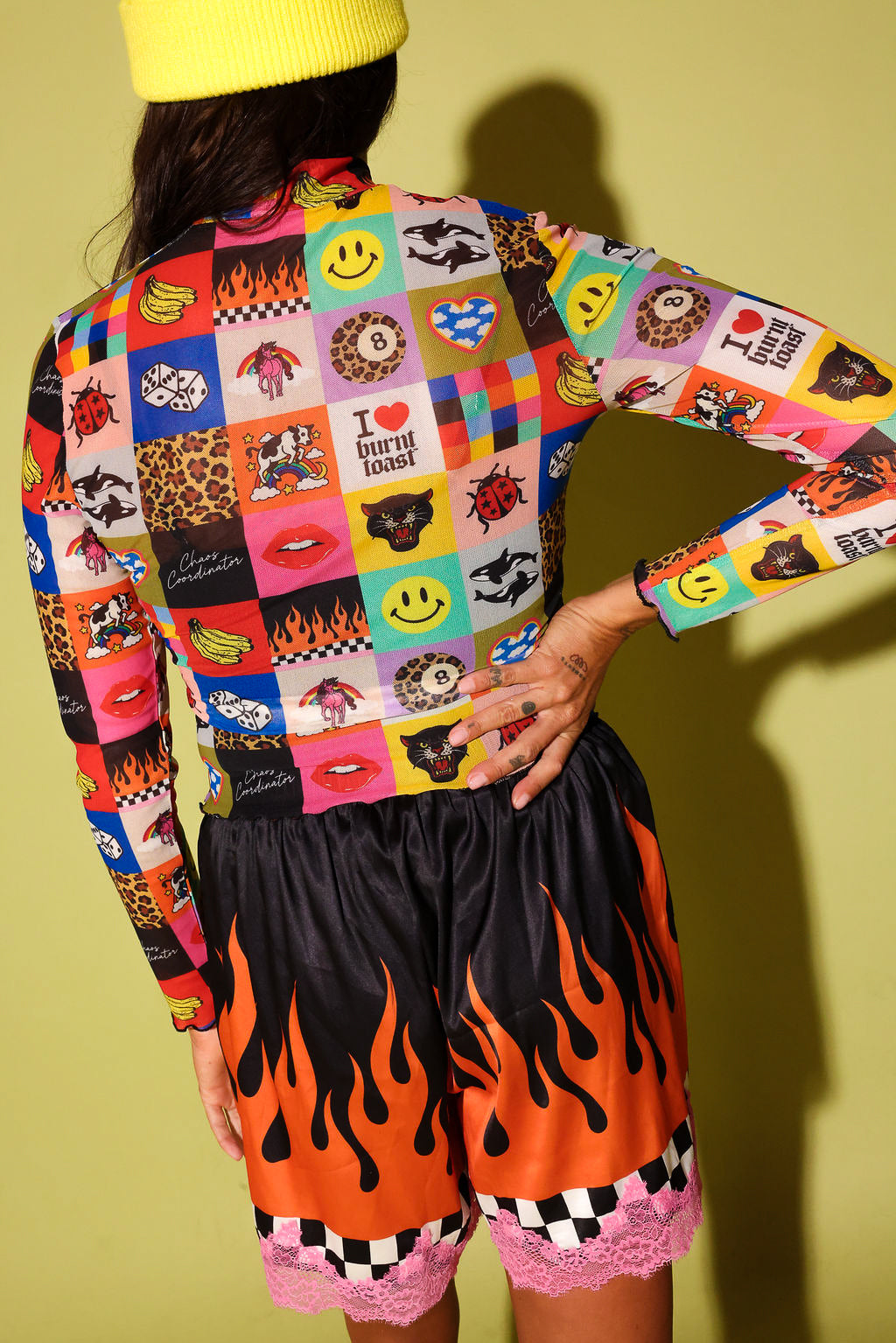 A model from behind wearing a vibrant, multicolor patchwork long-sleeve crop top and black shorts with a bright orange and black flame pattern, set against a solid olive green background.