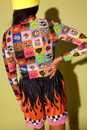 A model from behind wearing a vibrant, multicolor patchwork long-sleeve crop top and black shorts with a bright orange and black flame pattern, set against a solid olive green background.