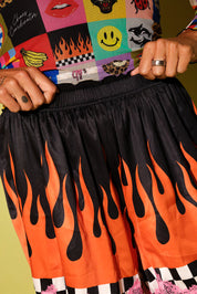 Black athletic shorts with a vibrant orange and black flame print, worn by a person holding the elastic waistband. The person is also wearing a colorful patchwork graphic t-shirt.