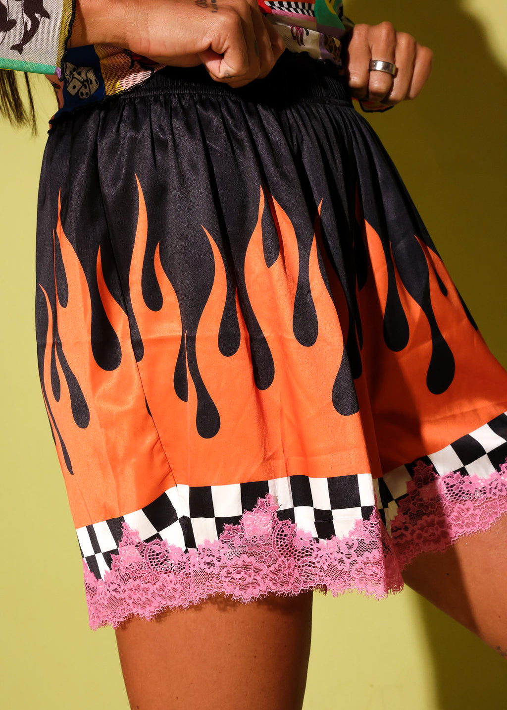 A close-up of a person wearing a short black skirt with a bold orange flame print, a black-and-white checkered band, and pink lace trim, posed against a yellow-green background.