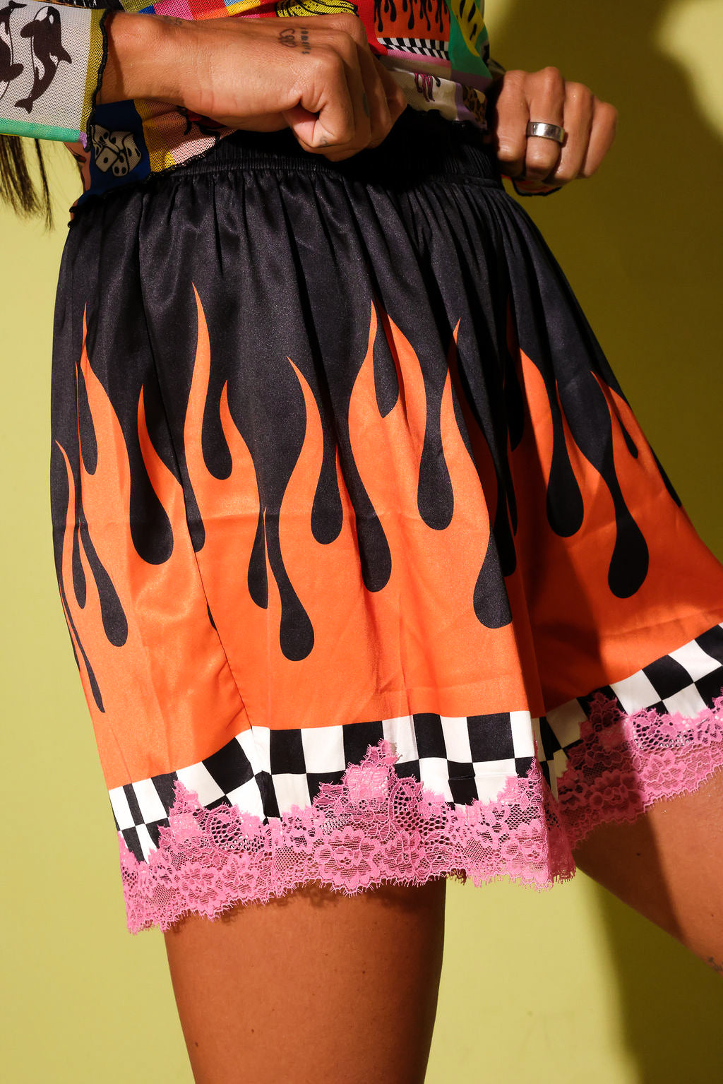 A close-up of a person wearing a short black skirt with a bold orange flame print, a black-and-white checkered band, and pink lace trim, posed against a yellow-green background.