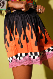 A close-up of a person wearing a short black skirt with a bold orange flame print, a black-and-white checkered band, and pink lace trim, posed against a yellow-green background.