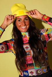 A smiling woman wearing a bright yellow knit beanie and a colorful patchwork long-sleeve top, posing against a solid pale yellow background.