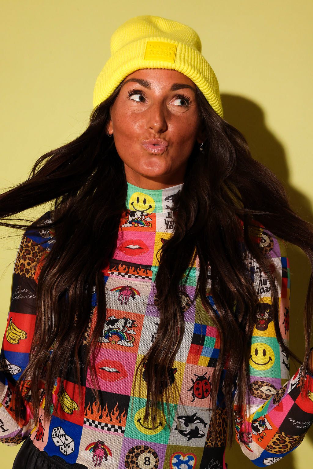 A woman wearing a bright yellow knit beanie and a colorful patchwork sweatshirt with smiley faces, lips, and leopard print, posing against a solid pale yellow background.