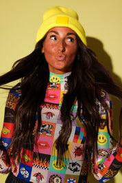 A woman wearing a bright yellow knit beanie and a colorful patchwork sweatshirt with smiley faces, lips, and leopard print, posing against a solid pale yellow background.