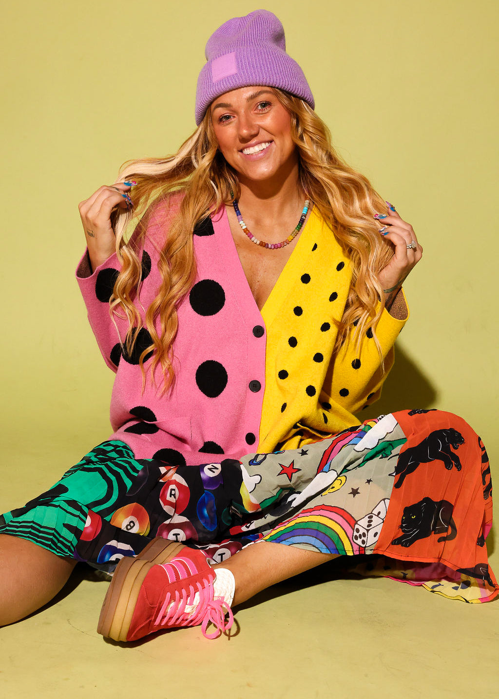 A woman sitting on a yellow-green studio background wearing a bright multicolor outfit with a pink and yellow polka-dot cardigan, a purple beanie, a rainbow-print skirt, and pink sneakers.