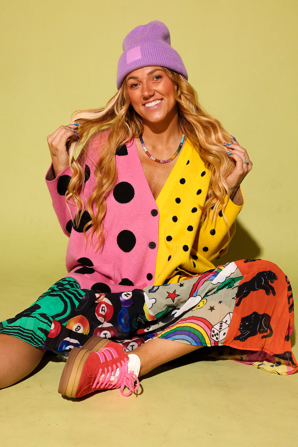 A woman sitting on a yellow-green studio background wearing a bright multicolor outfit with a pink and yellow polka-dot cardigan, a purple beanie, a rainbow-print skirt, and pink sneakers.