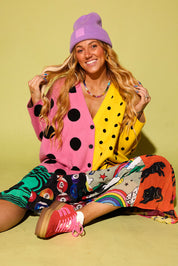 A woman sitting on a yellow-green studio background wearing a bright multicolor outfit with a pink and yellow polka-dot cardigan, a purple beanie, a rainbow-print skirt, and pink sneakers.