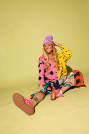 A woman sits on a pale yellow studio floor wearing a bright multicolor outfit with a pink and yellow polka-dot cardigan, a purple beanie, a colorful patterned skirt, and pink sneakers.