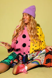 A woman sitting on a pale yellow-green studio background wearing a bright multicolor outfit with a pink and yellow polka-dot cardigan, patterned shorts, a purple beanie, and pink sneakers.