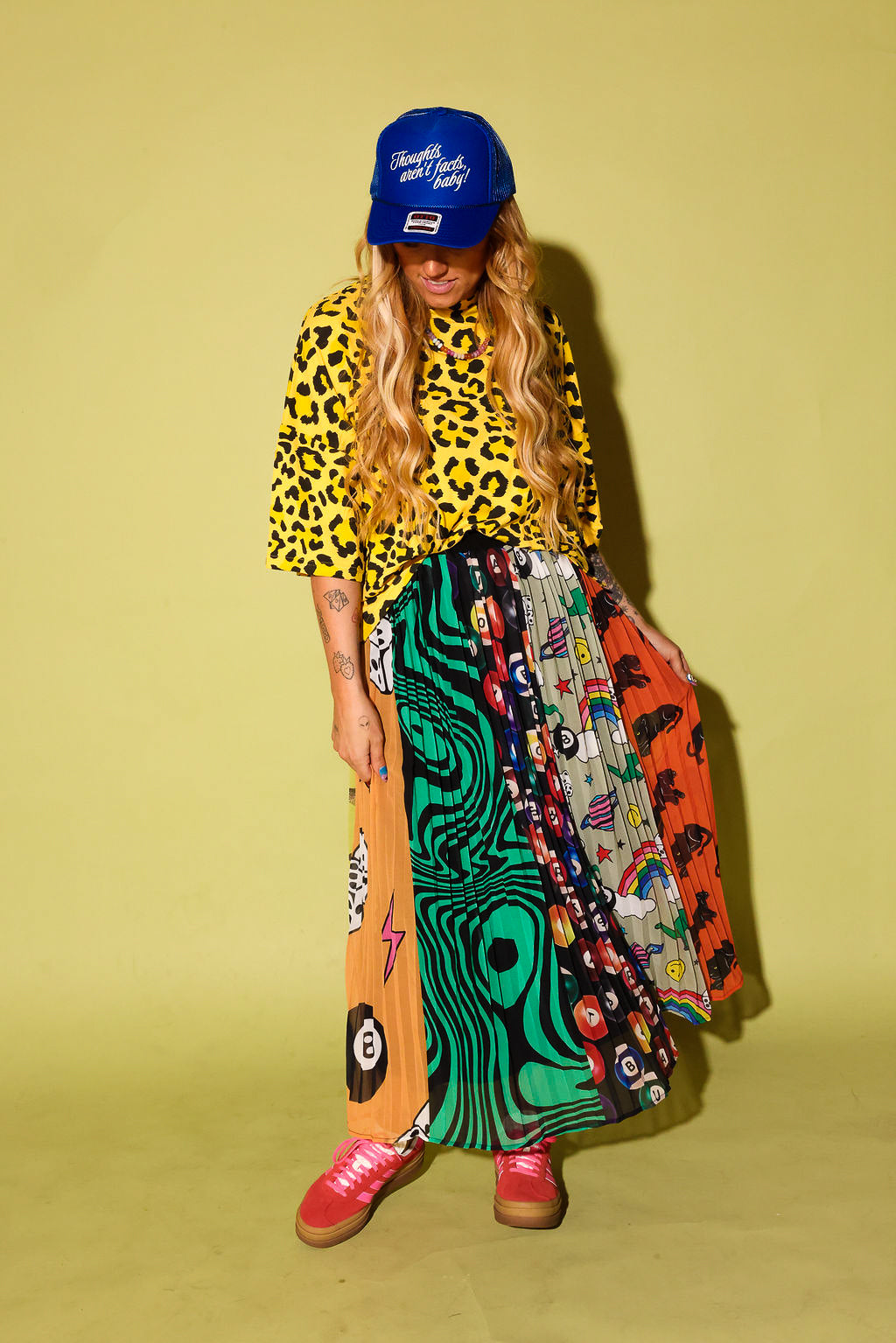 Model wearing a black and yellow leopard top, a plisse skirt with several different patterns and pink adidas, and a blue trucker hat with text on it, sitting against a pale yellow background