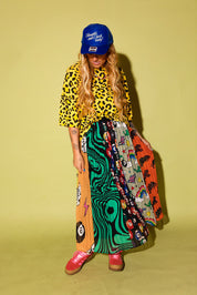 Model wearing a black and yellow leopard top, a plisse skirt with several different patterns and pink adidas, and a blue trucker hat with text on it, sitting against a pale yellow background