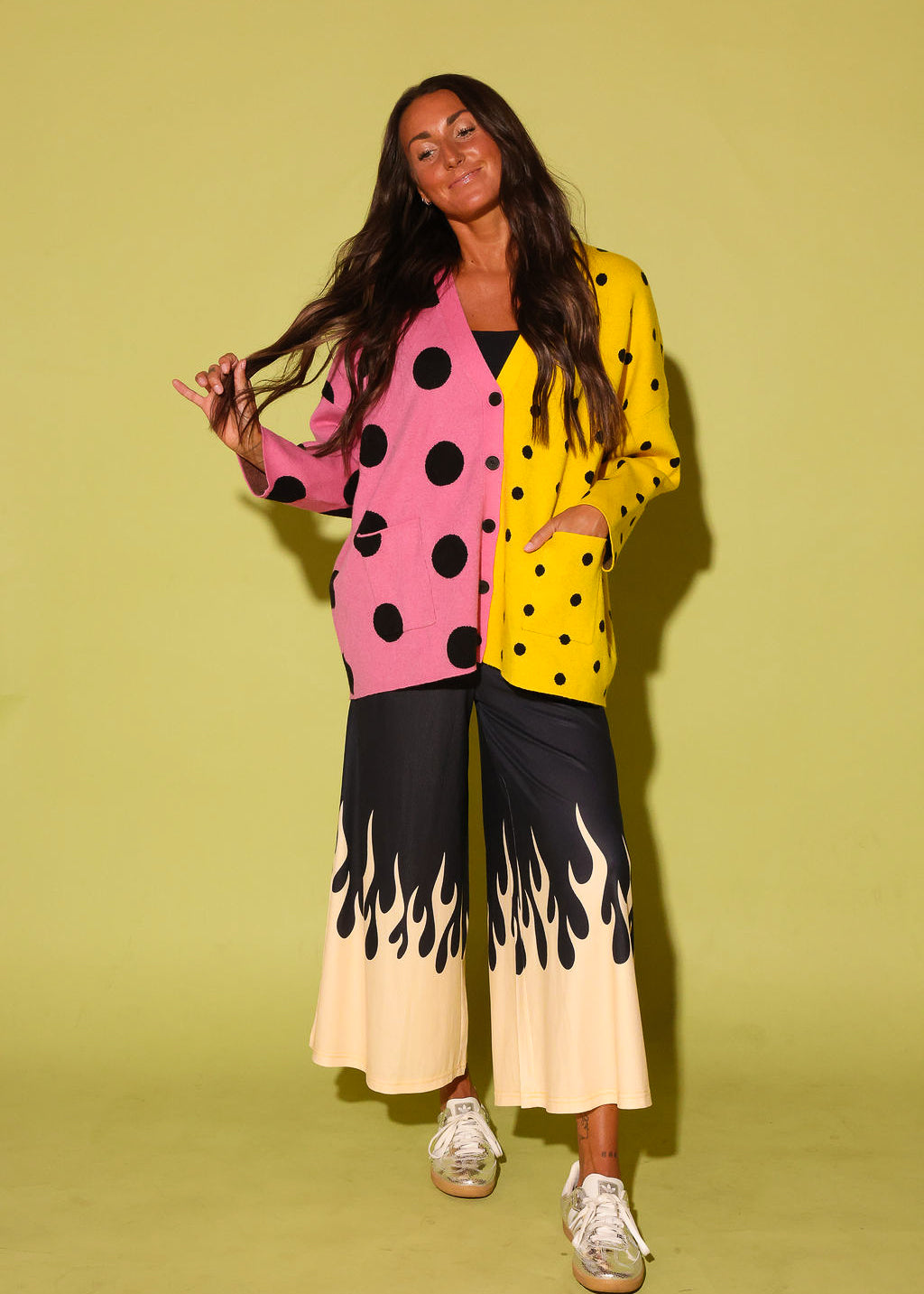 A woman modeling a colorful, split-design outfit consisting of a pink and yellow polka-dot cardigan, black and cream flame-print wide-leg pants, and white sneakers, set against a solid light yellow-green studio background.