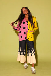 A woman modeling a colorful, split-design outfit consisting of a pink and yellow polka-dot cardigan, black and cream flame-print wide-leg pants, and white sneakers, set against a solid light yellow-green studio background.