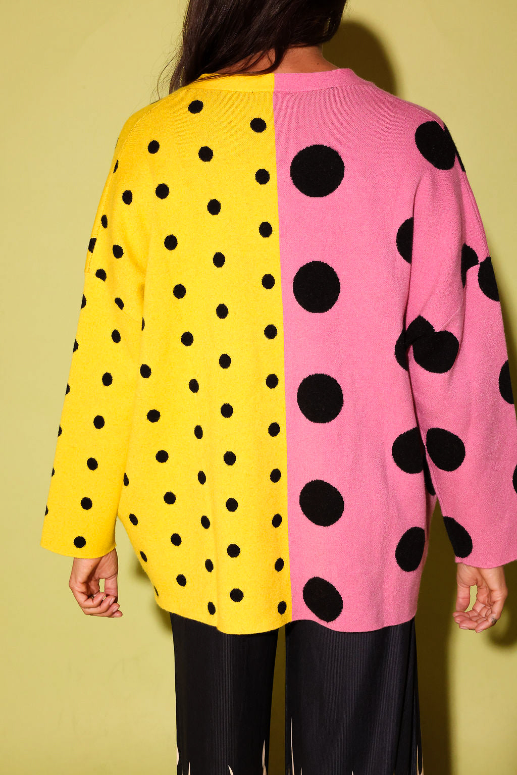 A person shown from the back wearing a long-sleeve knit top with a split design: one half is bright yellow with black polka dots and the other half is pink with large black polka dots, against a yellow background.