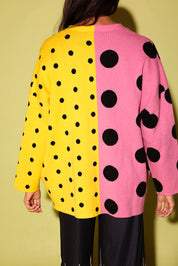 A person shown from the back wearing a long-sleeve knit top with a split design: one half is bright yellow with black polka dots and the other half is pink with large black polka dots, against a yellow background.