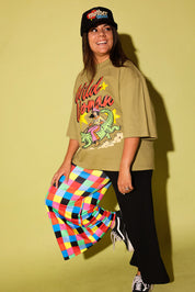 A woman posing in a vintage-style olive green graphic t-shirt, black pants, and a black cap with a colorful 'mother' logo, set against a solid yellow-green studio background.