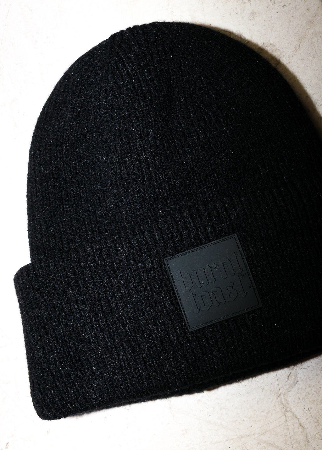 Close up of a black beanie with a burnt toast label on a white textured background