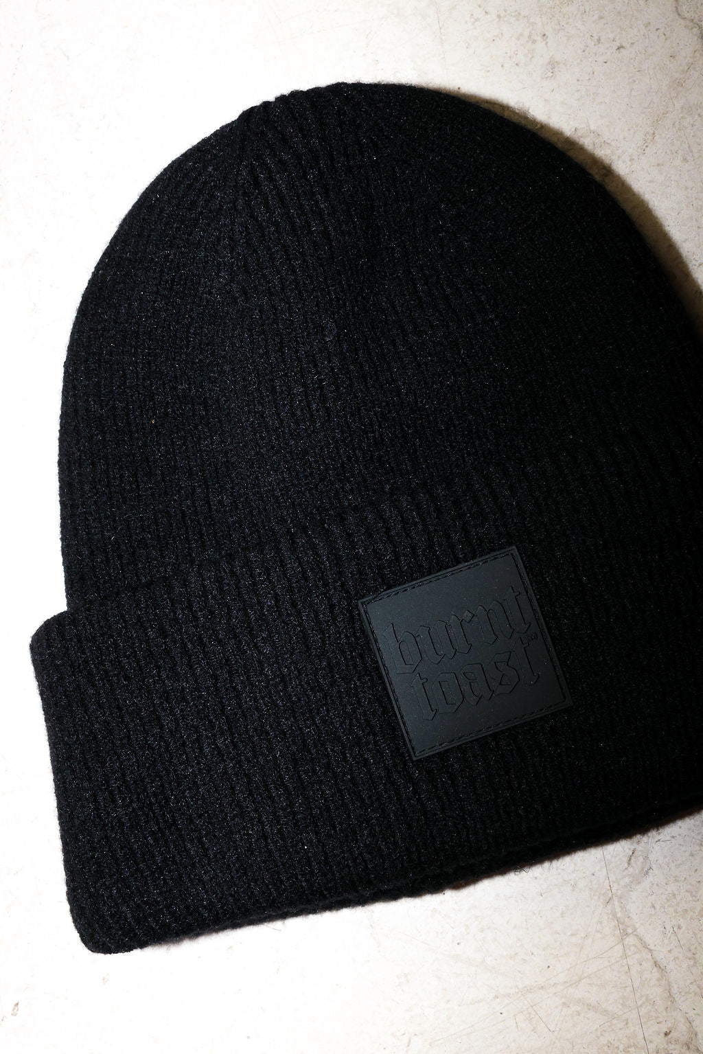 Close up of a black beanie with a burnt toast label on a white textured background