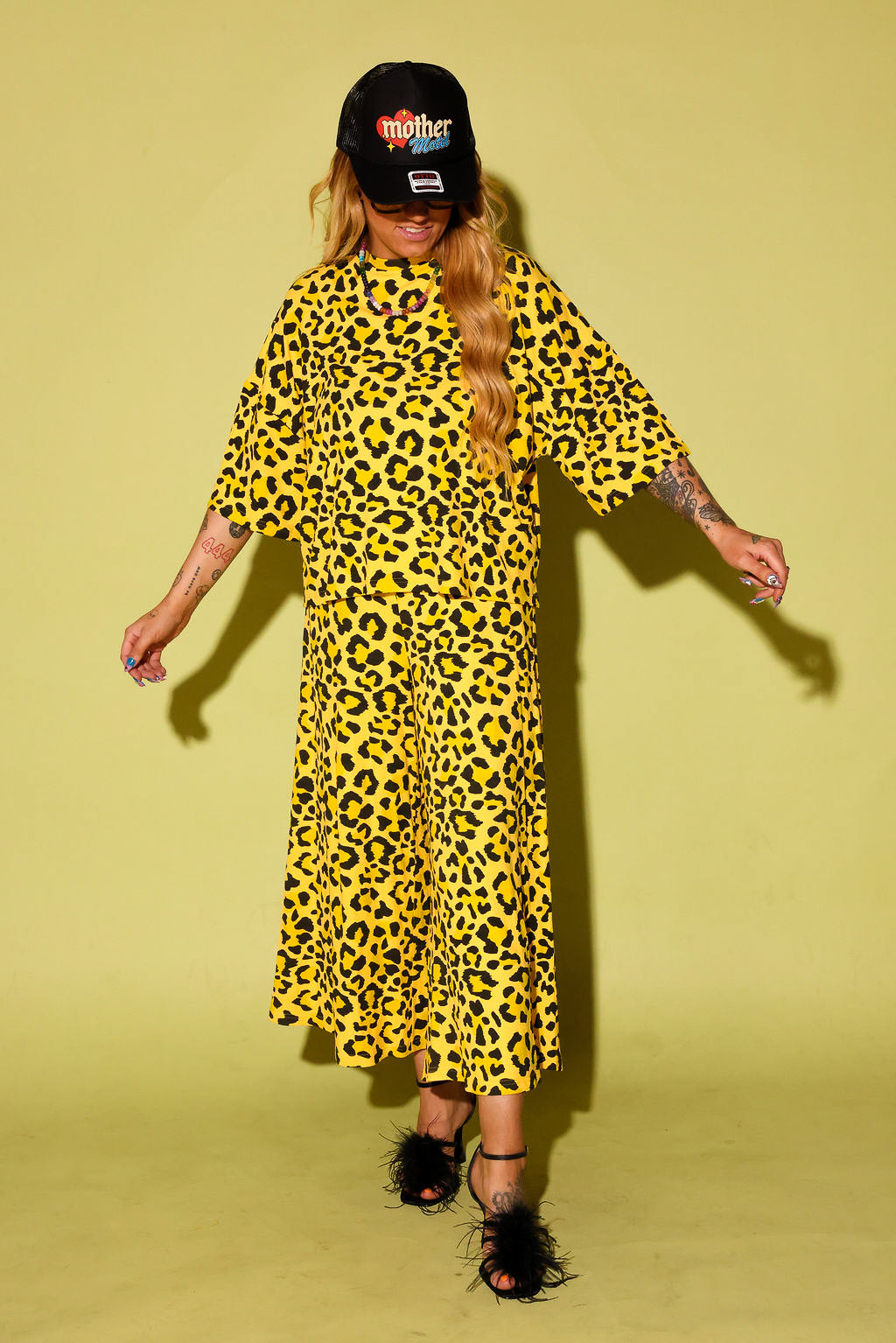 Model wearing a black and yellow leopard set black high heels and a blue trucker hat with text on it, sitting against a pale yellow background