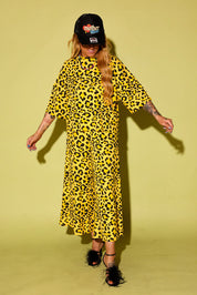 Model wearing a black and yellow leopard set black high heels and a blue trucker hat with text on it, sitting against a pale yellow background