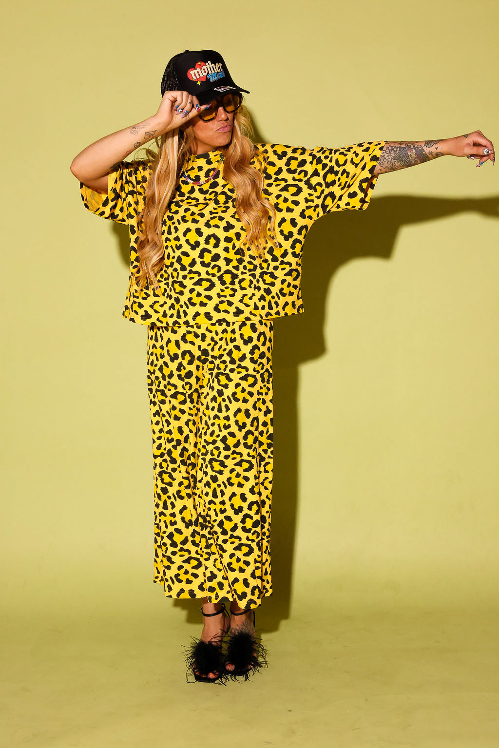 Model wearing a black and yellow leopard set black high heels and a black trucker hat that says 'mother' on it, against a pale yellow background