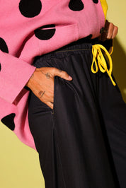 A close-up lifestyle shot of a person wearing black pinstriped wide-leg trousers with a yellow drawstring and a pink sweater with black polka dots, set against a solid pale yellow background.