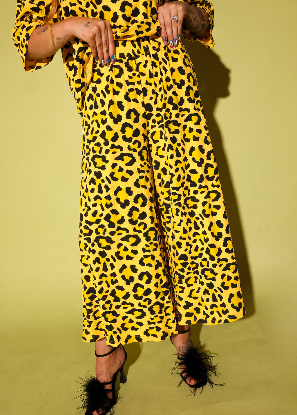 Model wearing a black and yellow leopard set black high heels holding the waistband, against a pale yellow background
