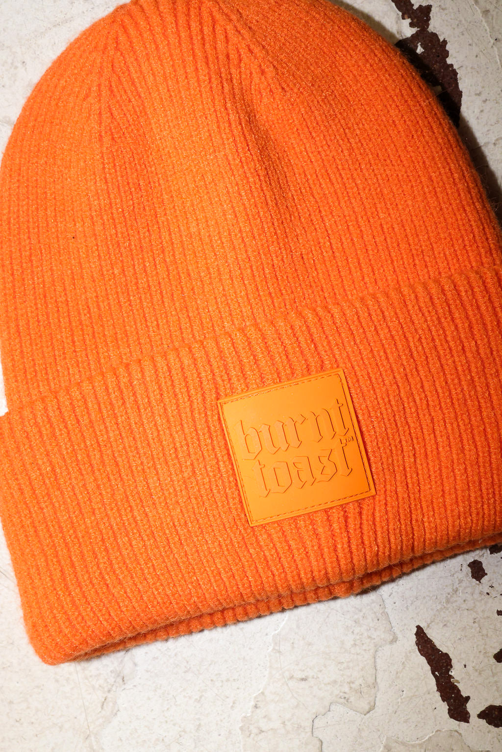 Close up of an orange beanie with a burnt toast label on a white textured background