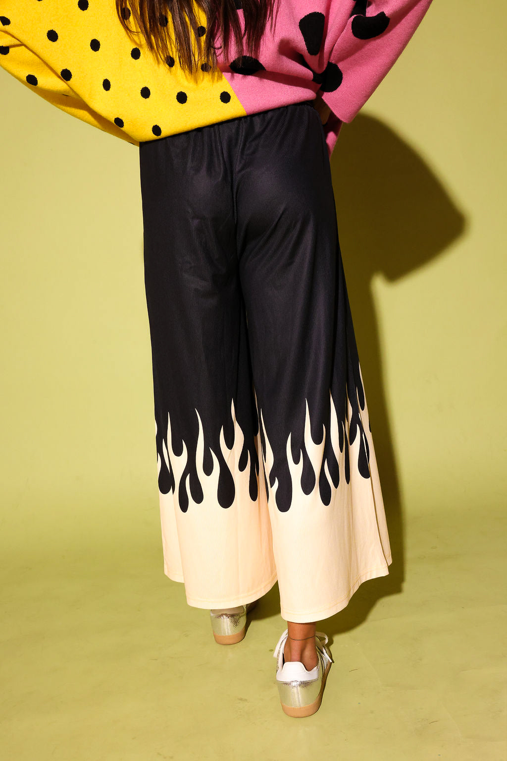 A person shown from the back wearing wide-leg cropped pants with a black upper section and a cream lower section with a black flame-like drip pattern at the hem, paired with a yellow and pink polka-dot top against a yellow-green studio background.