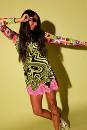 A woman modeling a long-sleeve mini dress with a black and lime green wavy abstract pattern, bright pink ruffle trim at the hem, and colorful cartoon patchwork sleeves, posed against a solid pale yellow-green studio background.