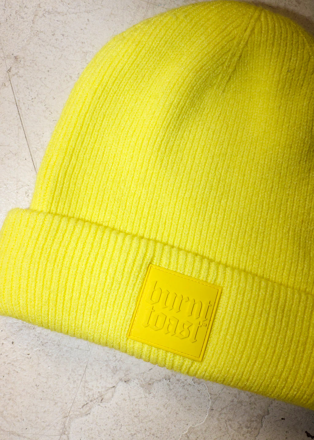 A bright neon yellow ribbed knit beanie hat with a matching yellow rectangular patch on the cuff that reads 'burn toast' in embossed lettering, shown on a light textured surface.