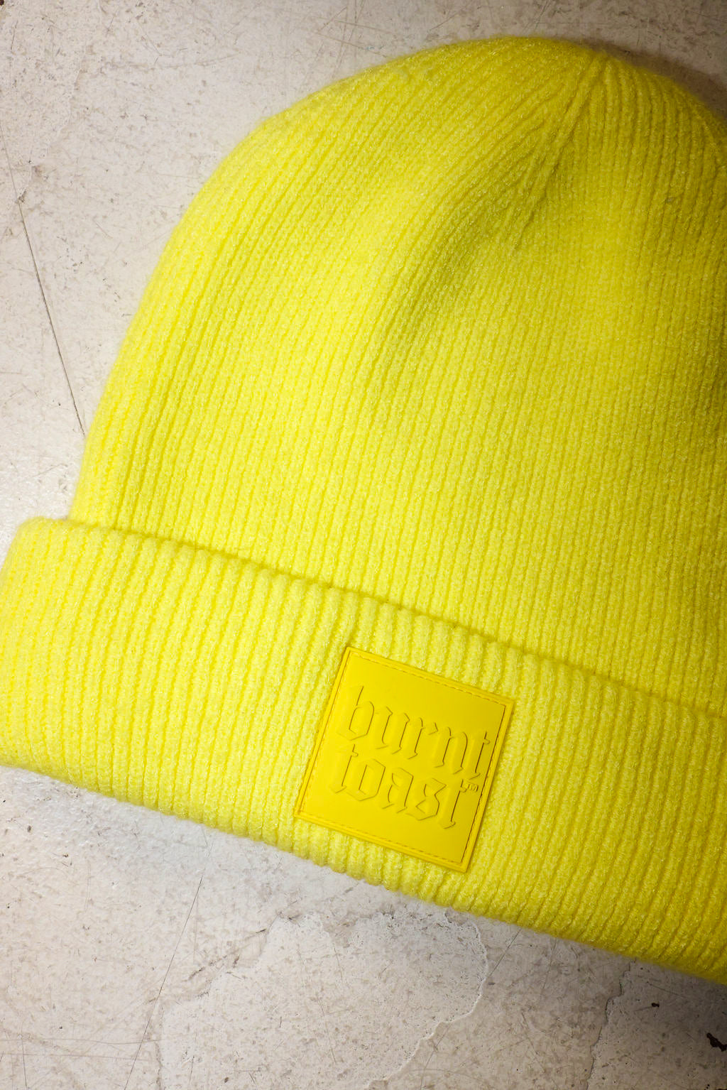 A bright neon yellow ribbed knit beanie hat with a matching yellow rectangular patch on the cuff that reads 'burn toast' in embossed lettering, shown on a light textured surface.