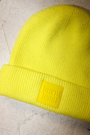 A bright neon yellow ribbed knit beanie hat with a matching yellow rectangular patch on the cuff that reads 'burn toast' in embossed lettering, shown on a light textured surface.