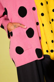A close-up of a person wearing a pink and yellow color-blocked cardigan with large black polka dots, paired with black pleated trousers. The person's hand is in their pocket.