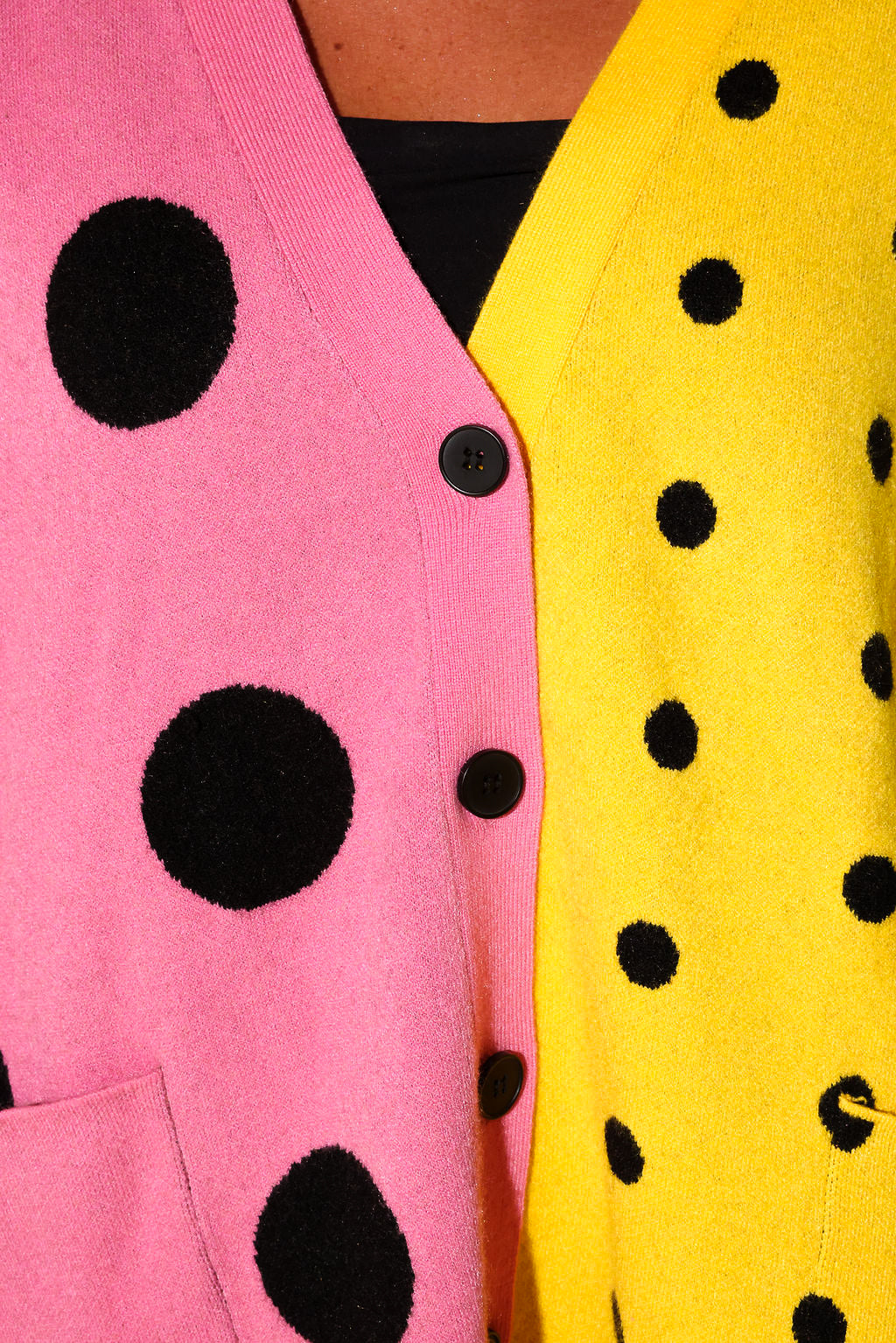 Close-up of a colorful cardigan with a split design: one side is pink with large black polka dots, and the other side is bright yellow with smaller black polka dots. A black button is visible at the center.