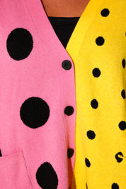 Close-up of a colorful cardigan with a split design: one side is pink with large black polka dots, and the other side is bright yellow with smaller black polka dots. A black button is visible at the center.