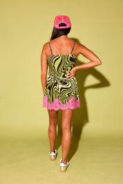 A woman is shown from behind wearing a short spaghetti-strap dress with a black and olive green wavy pattern and a pink ruffled hem, paired with a pink cap and white platform sandals, against a solid pale yellow-green studio background.