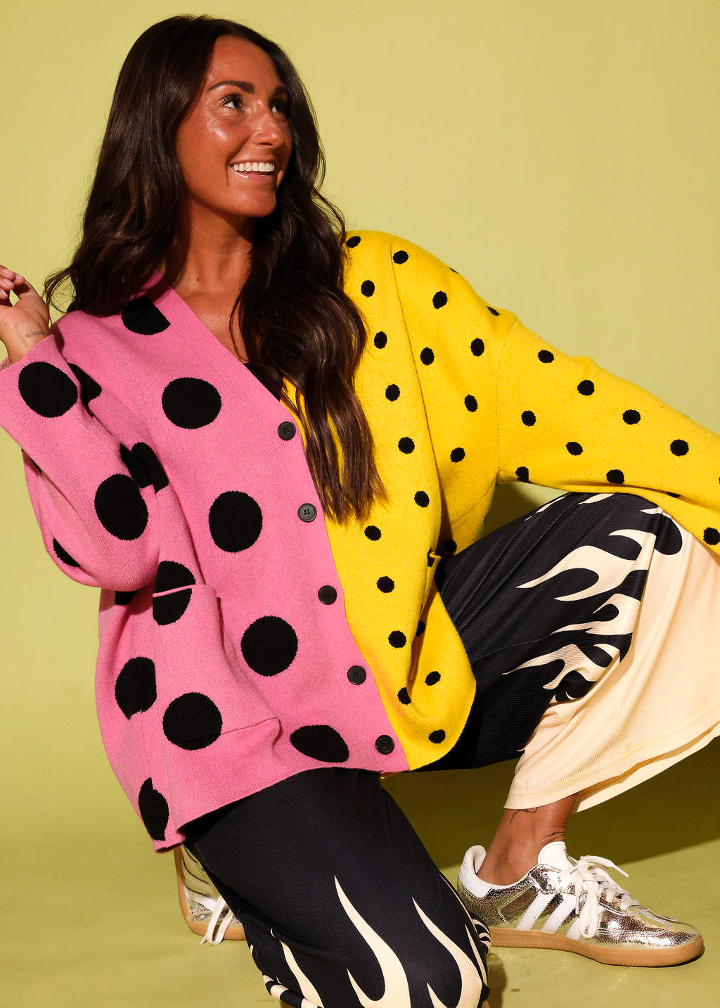 A woman crouches against a solid pale yellow-green studio background wearing a colorful cardigan with a pink and yellow color-blocked design and black polka dots, paired with black pants featuring a cream flame pattern and metallic sneakers.