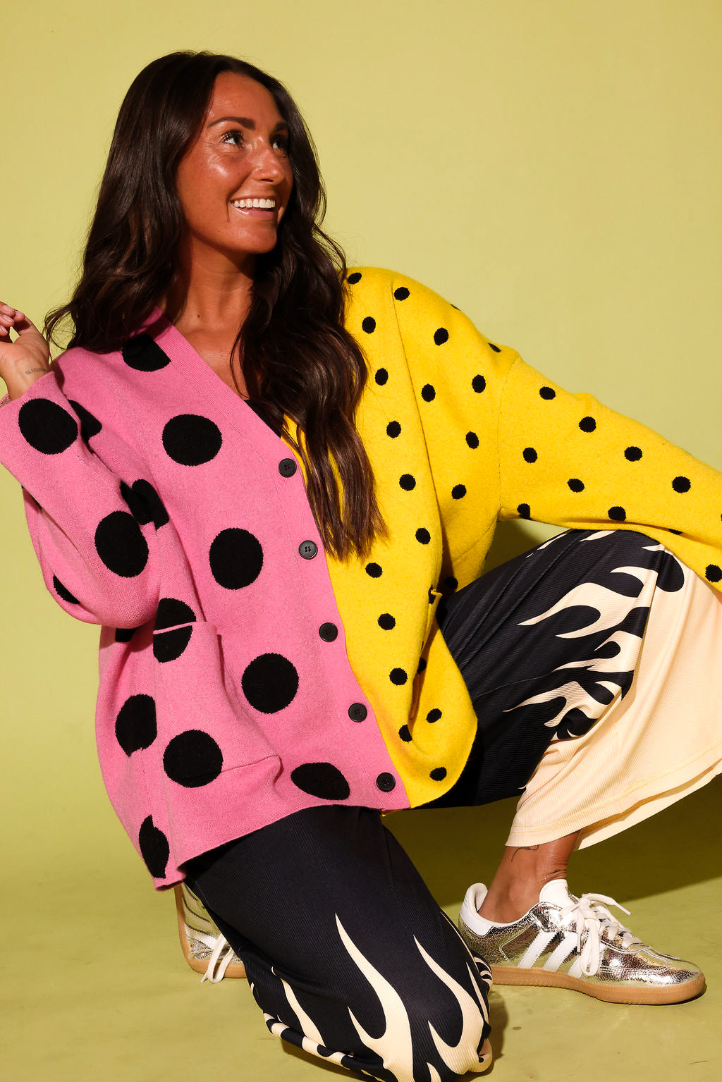 A woman crouches against a solid pale yellow-green studio background wearing a colorful cardigan with a pink and yellow color-blocked design and black polka dots, paired with black pants featuring a cream flame pattern and metallic sneakers.