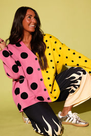 A woman crouches against a solid pale yellow-green studio background wearing a colorful cardigan with a pink and yellow color-blocked design and black polka dots, paired with black pants featuring a cream flame pattern and metallic sneakers.