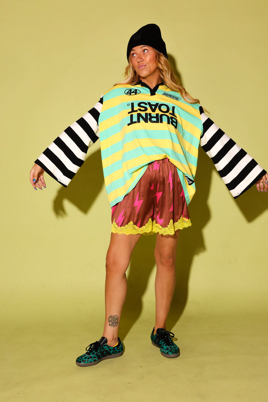 A model wearing a striped long sleeve polo with a Burnt Toast logo, maroon lace shorts with pink lightening bolts and yellow lace trim, a black beanie, and leopard adidas, against a pale yellow background