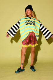 A model wearing a striped long sleeve polo with a Burnt Toast logo, maroon lace shorts with pink lightening bolts and yellow lace trim, a black beanie, and leopard adidas, against a pale yellow background