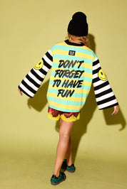 A model shown from behind wearing a colorful striped sweatshirt with the text 'DON'T FORGET TO HAVE FUN' on the back, paired with a black beanie, patterned shorts, and leopard print shoes, set against a solid yellow-green studio background.