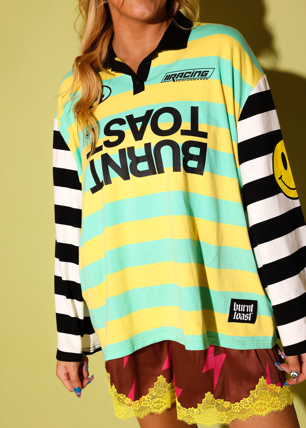 A woman wearing a colorful long-sleeve rugby-style shirt with yellow and mint green horizontal stripes, black-and-white striped sleeves, and bold black text reading 'BURNT TOAST' across the front. She is also wearing brown shorts with yellow lace and lightning-bolt details.