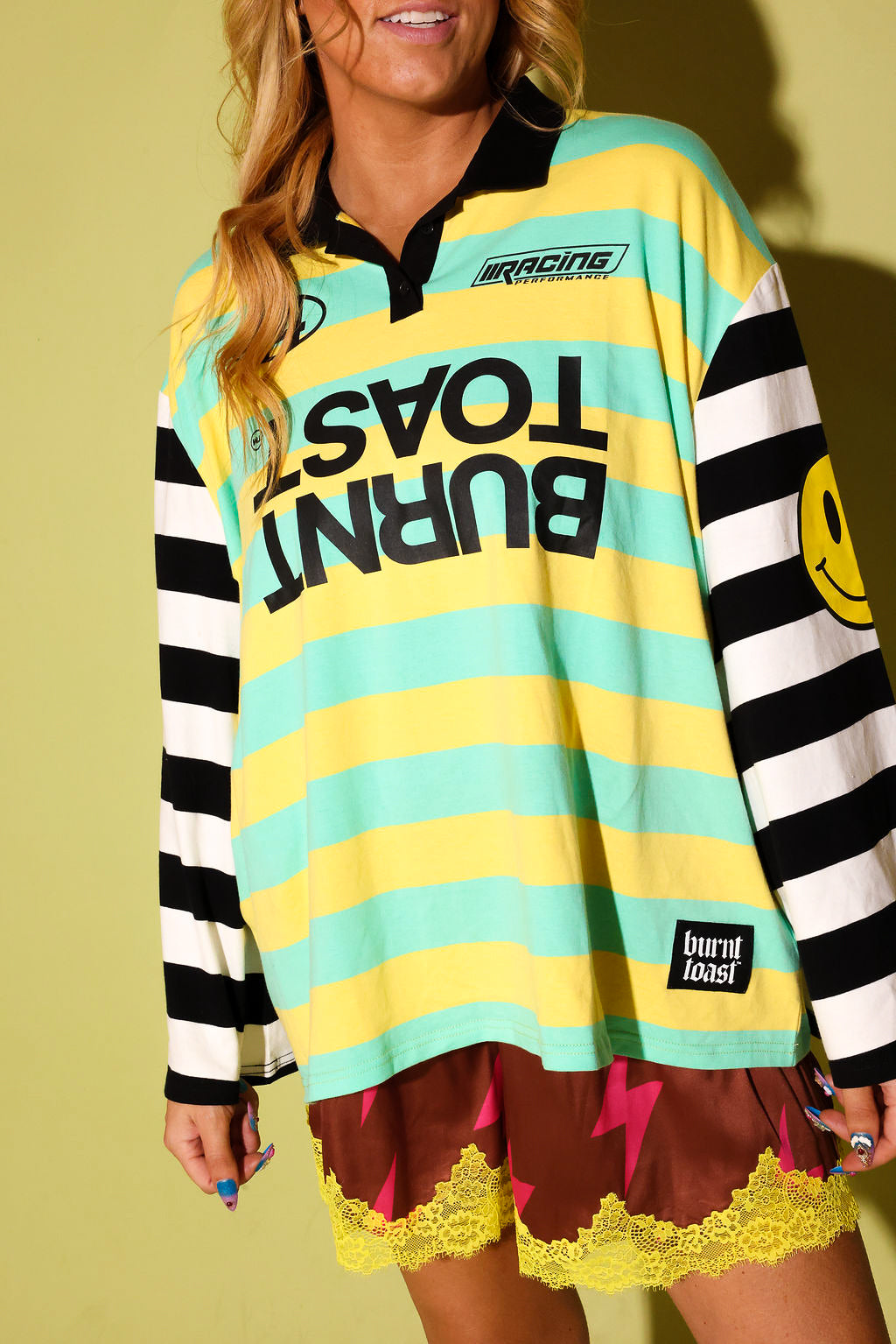 A woman wearing a colorful long-sleeve rugby-style shirt with yellow and mint green horizontal stripes, black-and-white striped sleeves, and bold black text reading 'BURNT TOAST' across the front. She is also wearing brown shorts with yellow lace and lightning-bolt details.