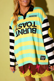 A woman wearing a colorful long-sleeve rugby-style shirt with yellow and mint green horizontal stripes, black-and-white striped sleeves, and bold black text reading 'BURNT TOAST' across the front. She is also wearing brown shorts with yellow lace and lightning-bolt details.