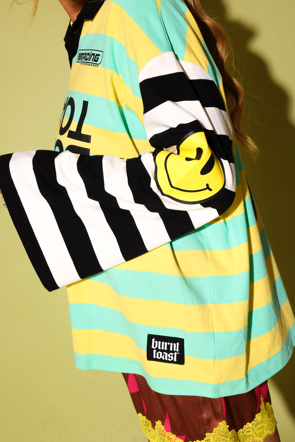 A close-up of a person wearing a colorful striped long-sleeve shirt with black and white stripes, yellow and mint green horizontal stripes, a yellow smiley face patch on the sleeve, and a 'burn! toast.' brand label near the hem.