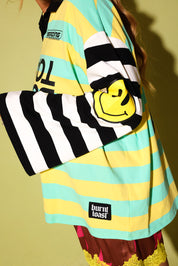 A close-up of a person wearing a colorful striped long-sleeve shirt with black and white stripes, yellow and mint green horizontal stripes, a yellow smiley face patch on the sleeve, and a 'burn! toast.' brand label near the hem.